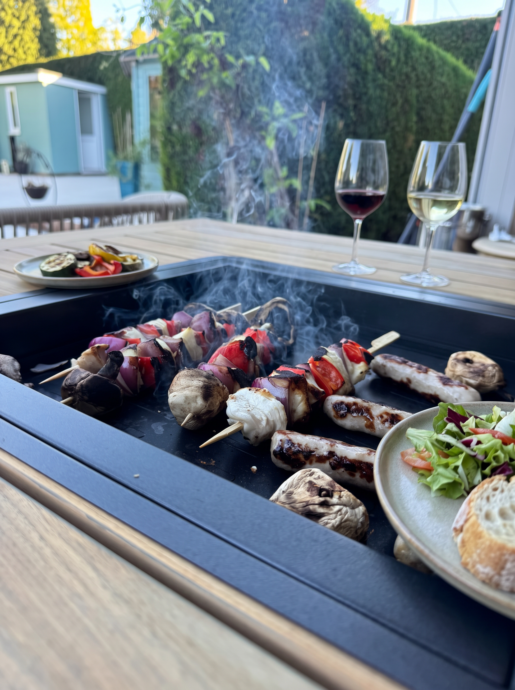 Food cooking on an Indugia teak grill table with plates and wine ready to share